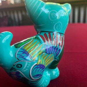 May include: A teal blue ceramic figurine of a cat with colorful painted details, including gold glitter accents. The cat is sitting with its tail curled around its body.