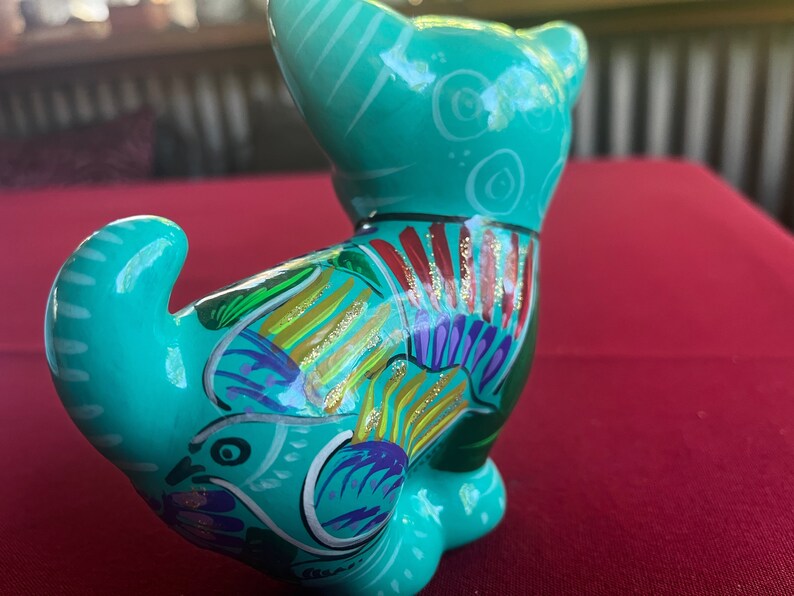 May include: A teal blue ceramic figurine of a cat with colorful painted details, including gold glitter accents. The cat is sitting with its tail curled around its body.