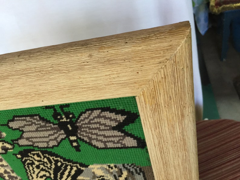 May include: A framed needlepoint picture with a green background and a butterfly design. The frame is made of wood and has a light brown finish.