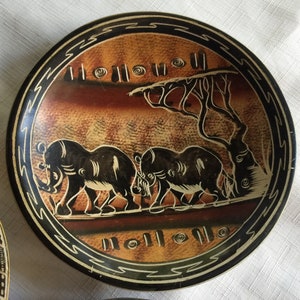 May include: A round, black and brown ceramic plate with a decorative design of three elephants walking in a line. The elephants are black and the background is brown with a black border.