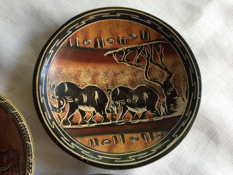 May include: A round, black and brown ceramic plate with a decorative design of three elephants walking in a line. The elephants are black and the background is brown with a black border.