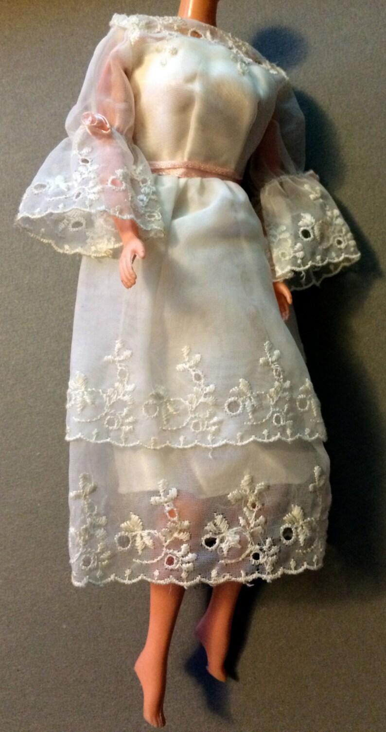Puede incluir: Un vestido blanco de encaje con una cinta rosa en la cintura. El vestido es de tela transparente y tiene bordados delicados.