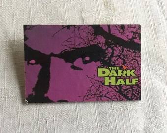 Op de afbeelding: Een paarse en zwarte poster met de tekst "THE DARK HALF" in groene letters. De poster toont een silhouet van een gezicht met een tak van een boom op de achtergrond.