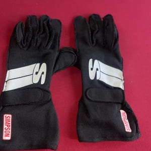 Puede incluir: Un par de guantes de carreras negros con detalles blancos y la letra "S" en el dorso. La marca "SIMPSON" es visible en la mu&ntilde;eca. Estos guantes est&aacute;n dise&ntilde;ados para deportes de motor.