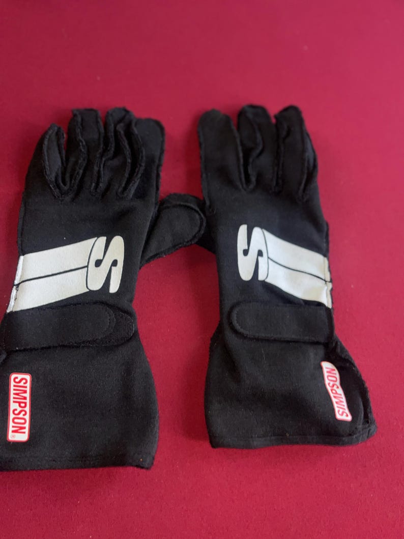 Puede incluir: Un par de guantes de carreras negros con detalles blancos y la letra "S" en el dorso. La marca "SIMPSON" es visible en la mu&ntilde;eca. Estos guantes est&aacute;n dise&ntilde;ados para deportes de motor.