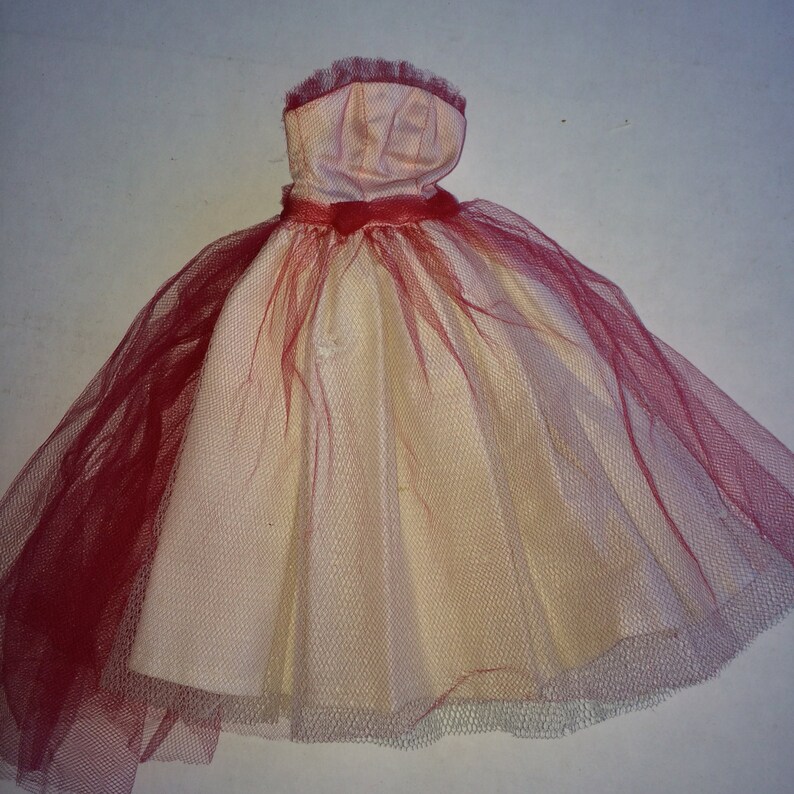May include: A doll-sized dress with a strapless bodice and a full skirt. The dress is primarily cream-colored, with a red tulle overlay on the skirt and a red ribbon at the waist. The bodice is a light pink color. The dress is designed for a doll.