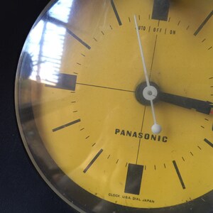 May include: A yellow clock face with black markings and a black hand pointing to the 10. The clock face says "Panasonic" and "Clock U.S.A. Dial Japan".