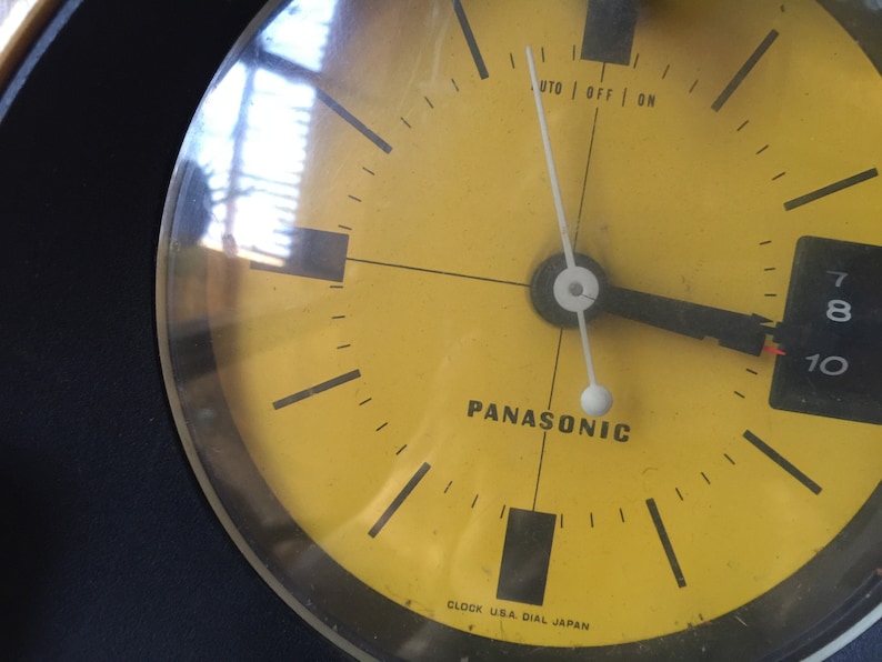 May include: A yellow clock face with black markings and a black hand pointing to the 10. The clock face says "Panasonic" and "Clock U.S.A. Dial Japan".