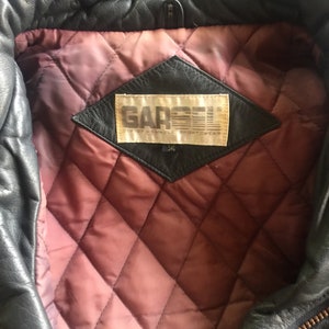 Pu&ograve; includere: Primo piano dell'interno di una giacca di pelle nera. La fodera &egrave; trapuntata e un'etichetta marrone con il testo "Gar&ccedil;on City of Reliable Sportswear" &egrave; visibile. L'etichetta ha anche il numero "34" stampato.