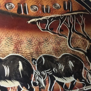May include: A close-up of a decorative plate with a black and white illustration of two rhinoceroses walking through a savanna landscape. The background is a brown and orange color with a white tree and white swirls.