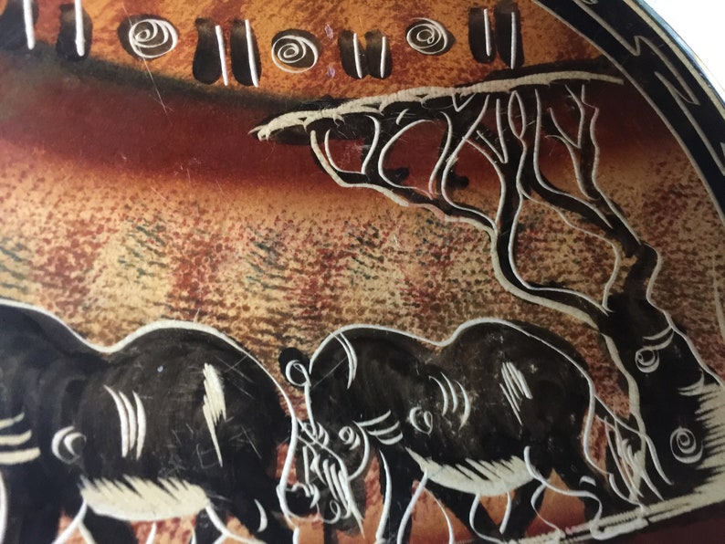 May include: A close-up of a decorative plate with a black and white illustration of two rhinoceroses walking through a savanna landscape. The background is a brown and orange color with a white tree and white swirls.