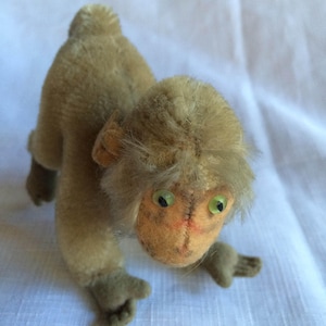 May include: A vintage plush monkey toy with brown fur, green eyes, and a light brown face. The monkey is in a crouching position with its arms outstretched.