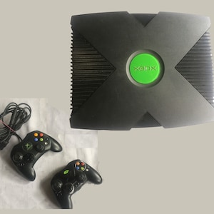 May include: A black Xbox console with a green XBOX logo on the front. Two black controllers are included.