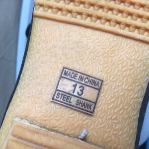 May include: Close-up of the bottom of a shoe with a tan rubber sole. The sole has a textured pattern and a label that reads "Made in China" and "Steel Shank". The label also shows the size "13".