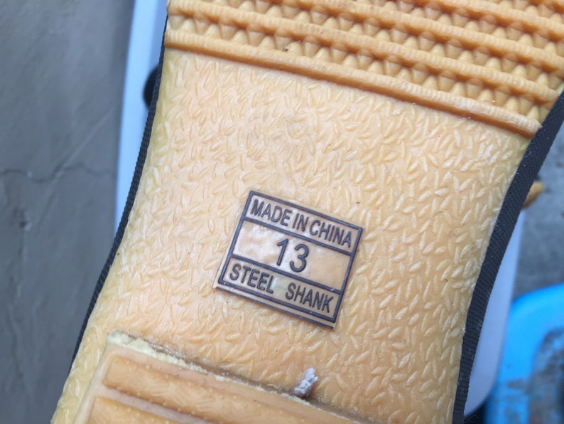 May include: Close-up of the bottom of a shoe with a tan rubber sole. The sole has a textured pattern and a label that reads "Made in China" and "Steel Shank". The label also shows the size "13".