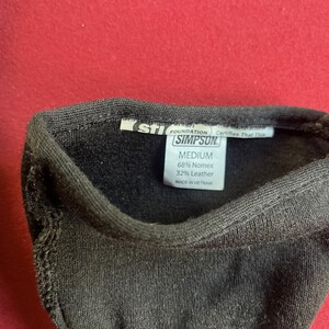 Puede incluir: Primer plano de un art&iacute;culo textil negro con una etiqueta que dice "SIMPSON" y "MEDIUM". La etiqueta tambi&eacute;n indica la composici&oacute;n del material: 68% Nomex y 32% cuero. El art&iacute;culo est&aacute; hecho en Vietnam.