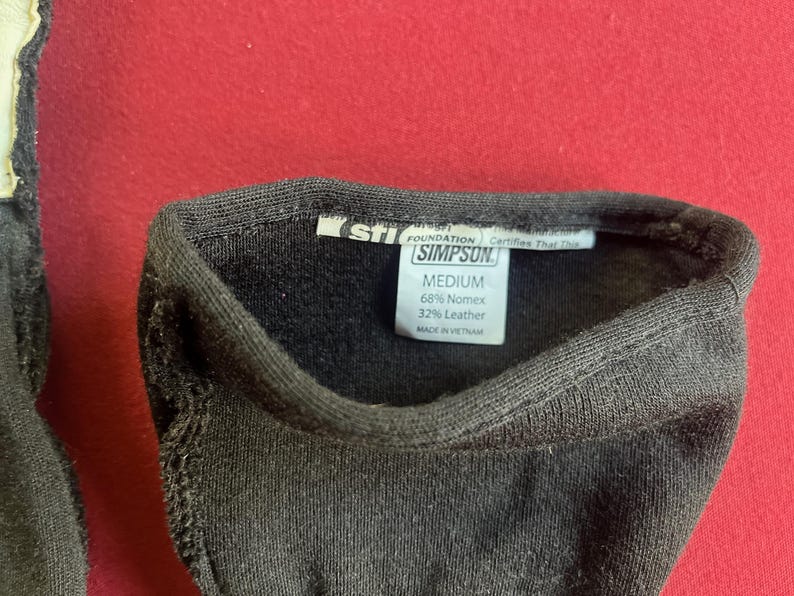 Puede incluir: Primer plano de un art&iacute;culo textil negro con una etiqueta que dice "SIMPSON" y "MEDIUM". La etiqueta tambi&eacute;n indica la composici&oacute;n del material: 68% Nomex y 32% cuero. El art&iacute;culo est&aacute; hecho en Vietnam.