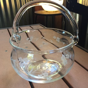 May include: A clear glass bowl with a handle, etched with a floral design. The bowl is sitting on a wooden surface.