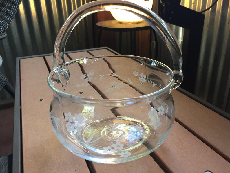 May include: A clear glass bowl with a handle, etched with a floral design. The bowl is sitting on a wooden surface.