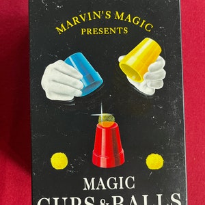 Könnte beinhalten: Marvin's Magic präsentiert Magic Cups & Balls. Eine schwarze Box mit einem grünen Kreis oben. Die Box zeigt eine weiße, behandschuhte Hand, die eine blaue Tasse hält, eine weiße, behandschuhte Hand, die eine gelbe Tasse hält, und eine rote Tasse mit einem gelben Ball darin.