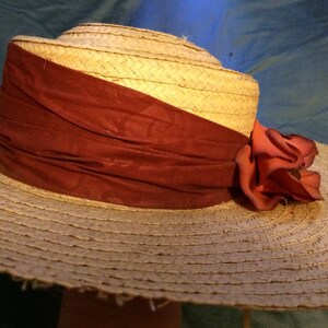 Pu&ograve; includere: Un cappello di paglia a tesa larga con un nastro color borgogna e un grande fiore color corallo.