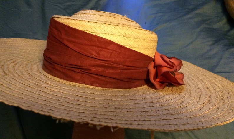 Pu&ograve; includere: Un cappello di paglia a tesa larga con un nastro color borgogna e un grande fiore color corallo.