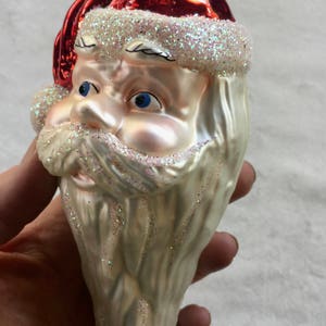 以下が含まれることがあります： サンタクロースの頭の形をしたクリスマスオーナメント。白いグリッターのトリムが付いた赤い帽子、青い目、グリッター付きの長い白いひげが特徴です。お祝いのホリデーデコレーション。