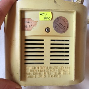 May include: A beige electric appliance with a metal grill and a warning label that reads "CAUTION: TO PREVENT ELECTRIC SHOCK DO NOT REMOVE COVER, NO USER SERVICEABLE PARTS INSIDE, REFER SERVICING TO QUALIFIED SERVICE PERSONNEL". The appliance has a serial number label that reads "SERIA № 91" and a UL sticker. A yellow sticky note with the text "42C/ RRSS" is attached to the appliance.