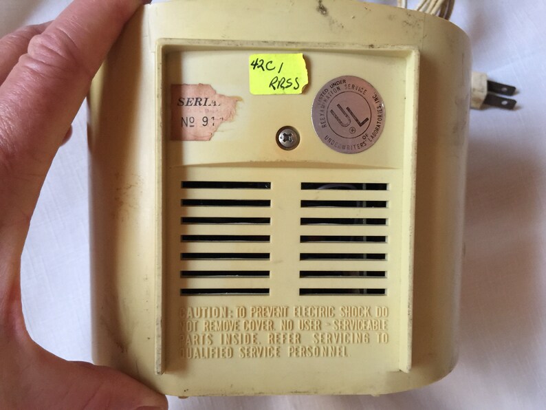 May include: A beige electric appliance with a metal grill and a warning label that reads "CAUTION: TO PREVENT ELECTRIC SHOCK DO NOT REMOVE COVER, NO USER SERVICEABLE PARTS INSIDE, REFER SERVICING TO QUALIFIED SERVICE PERSONNEL". The appliance has a serial number label that reads "SERIA № 91" and a UL sticker. A yellow sticky note with the text "42C/ RRSS" is attached to the appliance.