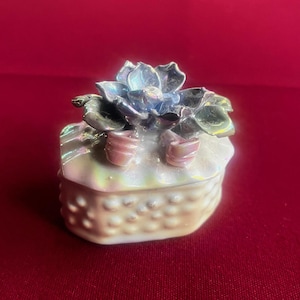 Peut inclure: Petite boîte à bijoux décorative avec une finition nacrée. Elle est ornée d'une fleur irisée bleue, verte et rose. La base présente un motif texturé et pointillé. Idéale pour ranger de petits objets.
