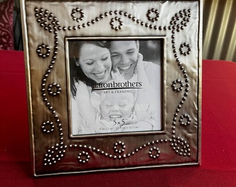 May include: A square metal photo frame with a distressed bronze finish. The frame is decorated with small, round, silver-colored studs and floral-like clusters. The frame holds a black and white photo and has the text "Aaron Brothers" and "5 x 5".