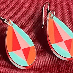 May include: A pair of teardrop-shaped earrings with a geometric design. The earrings are made of a light blue, pink, and orange material. The earrings have silver hooks.