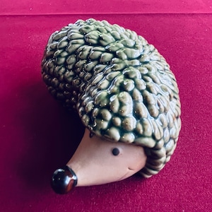 Op de afbeelding: Een keramieken egelbeeldje met een getextureerde, groen geglazuurde rug en een bruin gezicht. De egel heeft een donkerbruine neus en kleine zwarte ogen. Het beeldje staat tegen een dieprode achtergrond.