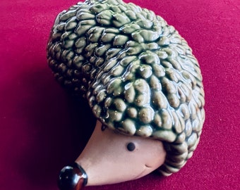 Puede incluir: Figura de erizo de cerámica con un dorso texturizado y esmaltado en verde y una cara de color canela. El erizo tiene una nariz marrón oscuro y pequeños ojos negros. La figura está sobre un fondo rojo oscuro.