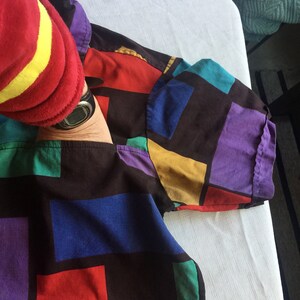 K&ouml;nnte beinhalten: Ein schwarzes, kurz&auml;rmeliges Hemd mit einem geometrischen Muster in Rot, Blau, Gr&uuml;n, Gelb und Lila. Das Hemd ist gefaltet und liegt flach auf einer wei&szlig;en Oberfl&auml;che.