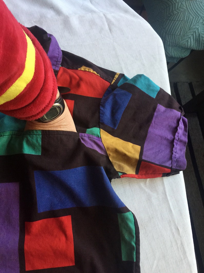 K&ouml;nnte beinhalten: Ein schwarzes, kurz&auml;rmeliges Hemd mit einem geometrischen Muster in Rot, Blau, Gr&uuml;n, Gelb und Lila. Das Hemd ist gefaltet und liegt flach auf einer wei&szlig;en Oberfl&auml;che.