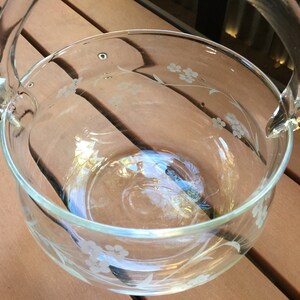 May include: A clear glass bowl with a floral design etched into the side. The bowl has two handles and is sitting on a wooden surface.