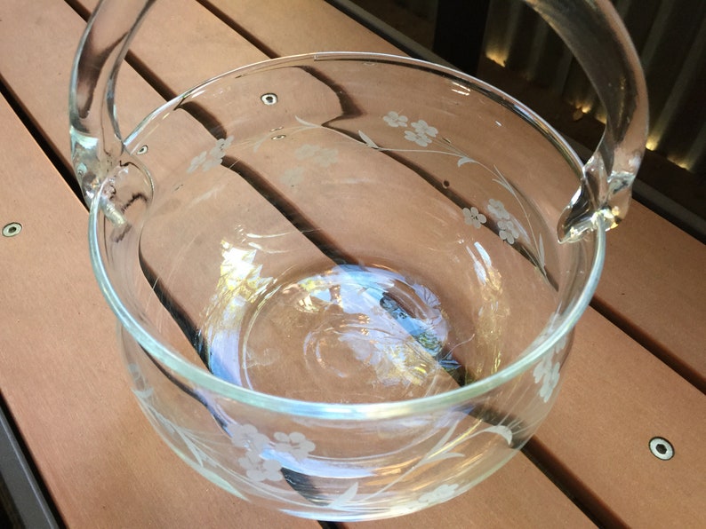 May include: A clear glass bowl with a floral design etched into the side. The bowl has two handles and is sitting on a wooden surface.