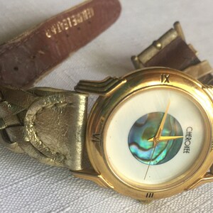 May include: Gold-tone wristwatch with a white face and a mother-of-pearl inlay. The watch has Roman numeral hour markers and a gold-tone band.