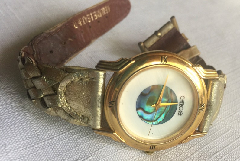 May include: Gold-tone wristwatch with a white face and a mother-of-pearl inlay. The watch has Roman numeral hour markers and a gold-tone band.