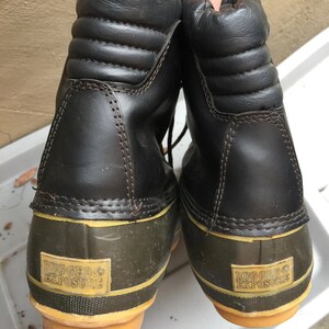 May include: A pair of black leather winter boots with a green rubber bottom and yellow trim. The boots have the words "Rugged Exposure" printed on the bottom.