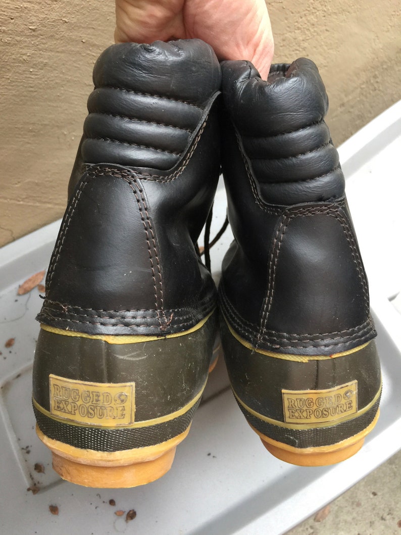 May include: A pair of black leather winter boots with a green rubber bottom and yellow trim. The boots have the words "Rugged Exposure" printed on the bottom.
