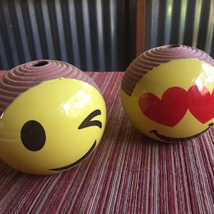 May include: Two ceramic round ornaments with brown tops and yellow smiley faces. One smiley face is winking and the other has red hearts for eyes.
