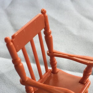 K&ouml;nnte beinhalten: Ein Miniatur-Schaukelstuhl aus Plastik, in rotbrauner Farbe lackiert. Der Stuhl hat eine hohe R&uuml;ckenlehne und ein schlichtes Design.