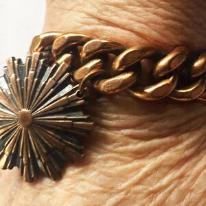 May include: A gold chain bracelet with a large, circular, sunburst-shaped pendant. The pendant is made of two-toned metal, with a darker center and lighter rays.