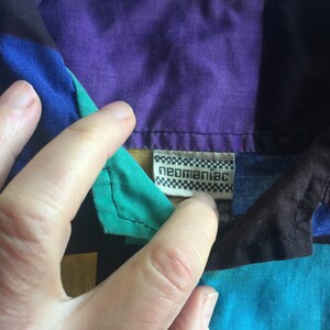 K&ouml;nnte beinhalten: Ein mehrfarbiges Hemd mit Kn&ouml;pfen und einem geometrischen Muster in Schwarz, Blau, Gr&uuml;n, Lila, Rot und Gelb. Das Hemd hat ein wei&szlig;es Etikett mit dem Text "neomaniac" in Schwarz.