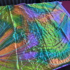 May include: A colorful, iridescent textile with a grid and curved line pattern. The fabric features a blend of green, orange, purple, and blue hues. The edges are trimmed with a solid purple border. The textile is likely a decorative item, such as a scarf or a throw.