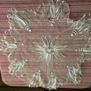 May include: A white crocheted doily with a delicate, lacy design. The doily has a circular shape and features intricate patterns throughout.