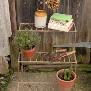 May include: A three-tiered, rusty metal plant stand with decorative arched detailing. The top shelf holds a ceramic vase with dried flowers and books. The middle shelf has gardening tools, and the bottom shelf has a potted plant. The stand is outdoors.