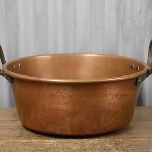 May include: A copper bowl with two arched handles. The bowl has a wide, shallow design and a rich, reddish-brown colour. The handles are made of a darker metal and are attached to the sides of the bowl. The bowl is sitting on a wooden surface.